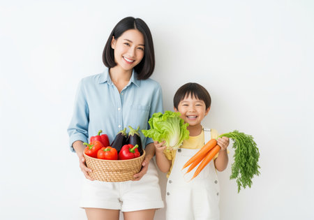 Smiling asian woman and young boy standing against a white background, proudly displaying a basket of fresh, colorful vegetables including carrots, lettuce, peppers, and eggplant, promoting healthy nutrition.の素材