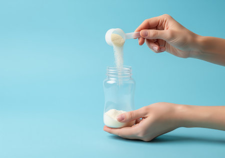 Woman hands preparing baby formula, pouring powdered milk from a scoop into a transparent feeding bottle against a bright blue background. essential infant nutrition and care concept.の素材