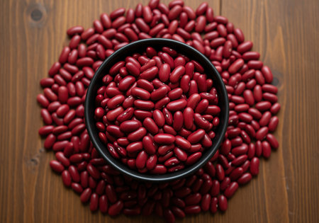 A close up overhead shot of dried red kidney beans piled high in a black bowl, with more beans scattered around the perimeter on a dark wood surface. ideal for food blogs, recipes, and healthy eating concepts.の素材