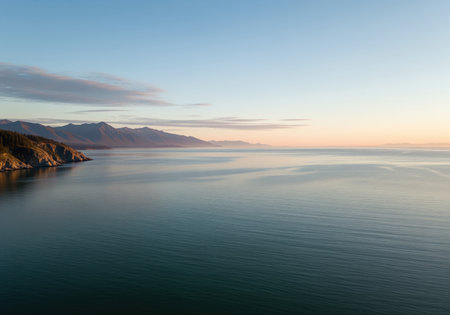 An expansive view of a calm ocean strait reflecting the soft colors of a sunrise sky. distant mountains frame the rocky, forested coastline, evoking peace and natural beauty.の素材
