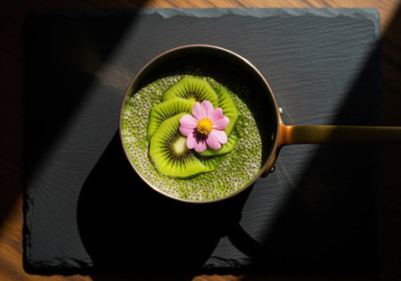 Vibrant green matcha chia pudding served in a small copper saucepan, garnished with fresh kiwi slices and a decorative pink flower on a dark slate board. healthy breakfast concept.の素材