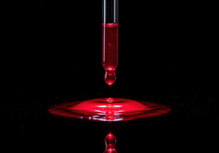 Red liquid drop suspended from a glass pipette, about to hit a black reflective surface, creating a dramatic splash and reflection. focus on science and experimentation.の素材