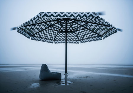 Oversized beach umbrella featuring a bold black and white chevron pattern standing next to a gray bean bag chair on a wide, wet, foggy beach. minimalist and moody scene.の素材