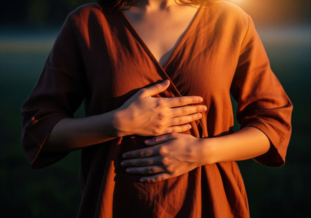Woman in a brown dress gently places her hands over her abdomen, illuminated by warm golden hour sunlight outdoors. focus on wellness, self care, body connection, and spirituality.の素材
