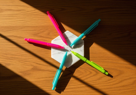 Vibrant pink, cyan, and green mechanical pencils arranged in a radial pattern, pointing toward the center of a white marble hexagon coaster, set on a warm wooden desk surface with dramatic sunlight and shadows.の素材