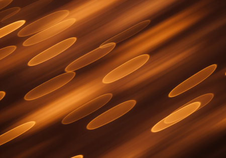 Abstract background featuring elongated, oval bokeh lights in warm amber and orange tones, creating a dynamic motion blur effect diagonally across the frame.の素材
