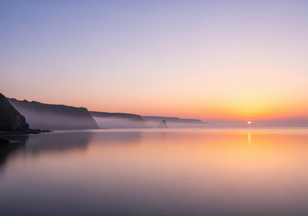 Serene coastal landscape featuring steep cliffs shrouded in morning mist, illuminated by a soft sunrise reflecting vibrant pastel colors across the perfectly calm water surface. tranquil nature scene.の素材