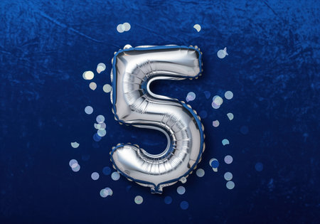 A shimmering silver foil balloon shaped like the number five is displayed against a rich, deep blue background, scattered with celebratory confetti. perfect for birthdays, anniversaries, or milestones.の素材