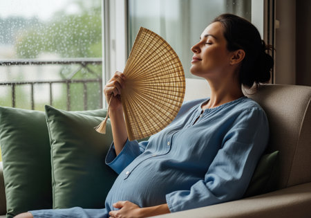 A pregnant woman sits on a sofa, using a woven hand fan to cool down and find relief from the heat and discomfort of pregnancy. she looks relaxed with her eyes closed.の素材