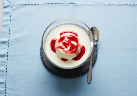 Panna cotta dessert topped with vibrant red berry sauce, served in a clear glass bowl on a dark slate coaster. overhead view on a light blue tablecloth with a spoon.の素材