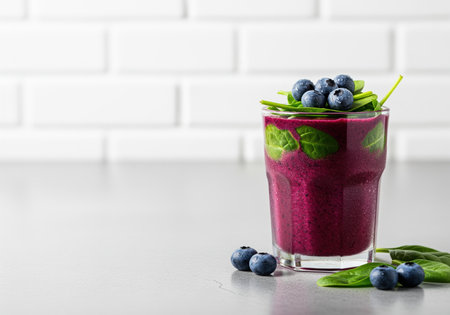 Frosty glass filled with a vibrant purple blueberry and spinach smoothie, garnished with fresh berries and green leaves, sitting on a gray kitchen counter with a white subway tile background. perfect for health, diet, and nutrition concepts.の素材