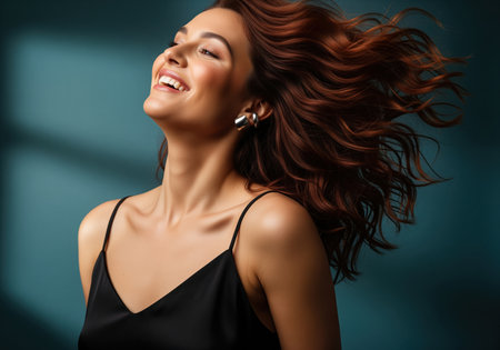 Young woman with voluminous auburn hair tossed in motion, smiling broadly and looking up. she wears a black silk top and silver earrings against a teal studio background, conveying happiness and beauty.の素材