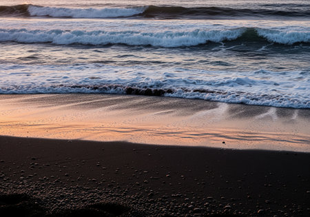 Powerful ocean waves cresting and breaking into white foam along a dark, volcanic black sand beach. the wet sand reflects the soft pink and orange hues of the twilight sky, emphasizing stark natural contrast and elemental power.の素材
