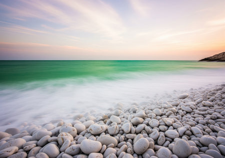 Smooth white pebble beach bordering the emerald turquoise sea under a pastel sunset sky. long exposure creates misty, ethereal waves washing ashore, symbolizing tranquility.の素材