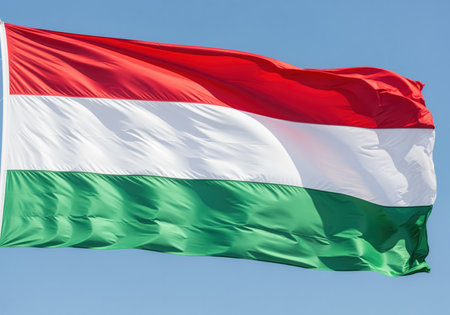 National flag of hungary featuring horizontal stripes of red, white, and green, fluttering strongly in the wind under a bright blue sky. symbolizing patriotism and national identity.の素材