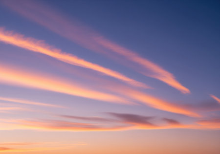 Long, parallel cirrus clouds illuminated by the setting sun, creating vibrant streaks of orange, peach, and pink against a deep blue and purple twilight sky gradient. peaceful and atmospheric nature background.の素材