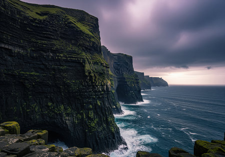 Towering vertical sea cliffs covered in dark rock and green moss overlooking the turbulent blue atlantic ocean. dramatic, moody sky creates a rugged nature landscape.の素材