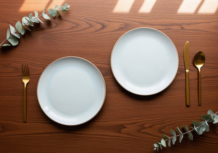 Refined table setting featuring two white dinner plates with gold rims, golden cutlery, and eucalyptus branches arranged on a dark walnut wood surface. ideal for menu mockups or upscale dining concepts.の素材