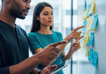 Diverse business colleagues analyzing project strategy and brainstorming ideas using colorful sticky notes on a whiteboard wall during a focused meeting.の素材