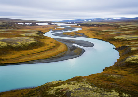 Winding turquoise glacial river meanders through the vast, colorful autumn tundra landscape, featuring mossy hills and distant snow patches under an overcast sky.の素材