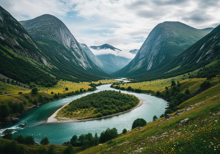 A wide turquoise river meanders through a lush green valley floor, surrounded by massive, steep, slate grey mountains under a cloudy sky. dramatic natural landscape scene featuring wilderness and geography.の素材