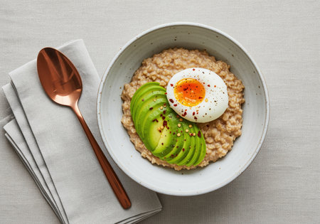 Savory oatmeal served in a white bowl, garnished with sliced avocado and a soft poached egg, sprinkled with chili flakes and pepper. healthy breakfast, top view.の素材