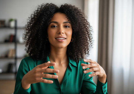 Professional young black woman with curly hair wearing a green blouse, actively gesturing and speaking directly to the camera during a virtual meeting or online presentation.の素材