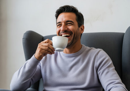 Hispanic man sitting comfortably in a gray armchair, laughing heartily while drinking from a white coffee cup. represents happiness, relaxation, and casual lifestyle.の素材