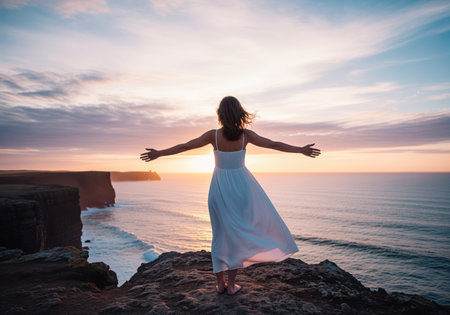Woman in a white dress standing on a rugged cliff edge with arms wide open, facing the vast ocean during a colorful sunset. concepts of freedom, travel, and adventure.の素材