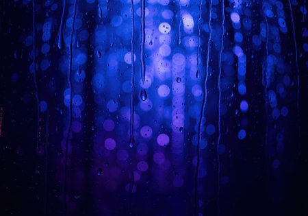 Raindrops and streaks of water flow down a dark window pane, obscuring abstract blue and purple bokeh lights from the city street at night. atmospheric background texture.の素材