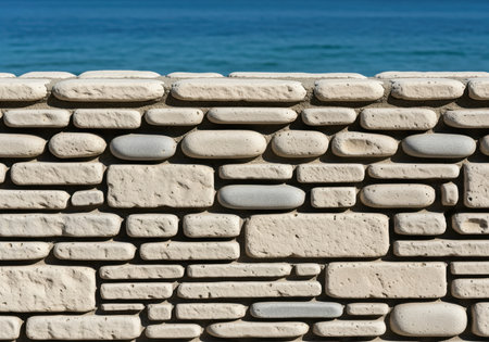 Beige natural stone wall texture composed of rounded river pebbles, set against a blurred background of bright blue ocean water. ideal for copy space or architectural detail.の素材