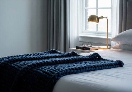 Luxurious modern bedroom interior featuring a white bed, deep navy chunky knit blanket, and a brass desk lamp illuminating a book and reading glasses on a nightstand near a window.の素材