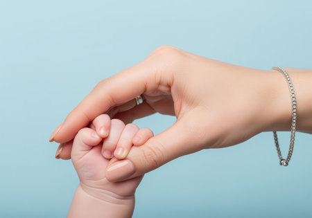 Adult woman hand gently holds the tiny curled fingers of a newborn baby against a light blue background, symbolizing protection, care, and the strong family bond between mother and child.の素材
