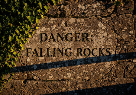 Weathered stone wall featuring an engraved warning sign reading danger falling rocks. textured surface covered in lichen and ivy, illuminated by dramatic sunlight, symbolizing risk and caution.の素材