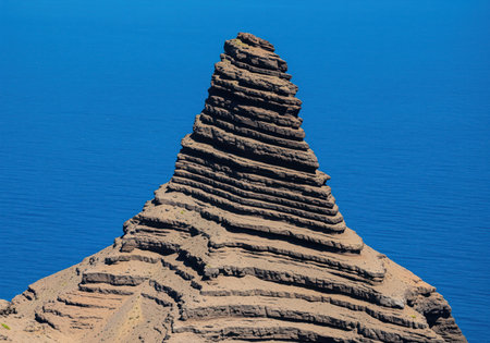 Rugged conical rock formation displaying prominent layered geological strata, contrasting sharply with the vast, deep blue ocean water below. natural erosion texture.の素材