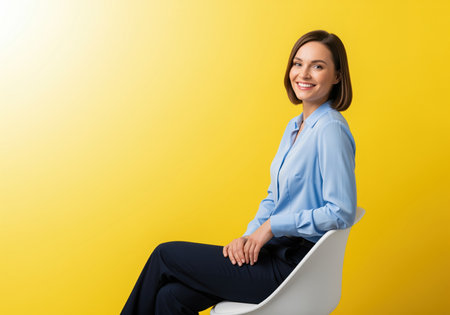 Professional young woman in a blue shirt and dark pants sitting on a modern white chair, smiling confidently against a vibrant yellow studio backdrop. this portrait conveys success and approachability.の素材