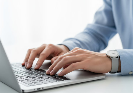 Professional hands wearing a blue shirt and wristwatch typing quickly on a modern silver laptop keyboard. focus on business, technology, communication, and efficiency in a bright office setting.の素材