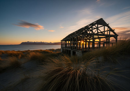 Skeletal wooden structure standing on coastal sand dunes, dramatically silhouetted by the setting sun. distant mountains and ocean visible under a vibrant sky.の素材