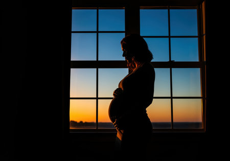 Pregnant woman standing in silhouette against a large window during sunset, gently touching her belly. the dark interior contrasts sharply with the vibrant blue and orange sky, symbolizing anticipation and new life.の素材