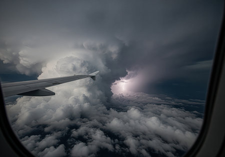 Airplane wing seen from the passenger window flying through dense, dark cumulonimbus storm clouds. a bright lightning strike is visible in the distance, symbolizing danger and extreme weather.の素材