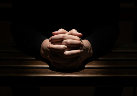 Clasped hands resting on a dark wooden church pew, illuminated by a single beam of light against a black background. represents prayer, faith, spirituality, grief, and contemplation in a dramatic, low key setting.の素材