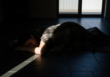 A woman lies covered by a plaid blanket on a dark tiled floor, resting or sleeping in deep shadow. a strong beam of sunlight cuts across the scene, highlighting the dramatic contrast.の素材