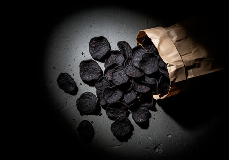 Dark, round black chips spill dramatically from a crumpled kraft paper bag onto a moody, textured surface under focused lighting. this savory snack food image is ideal for culinary projects.の素材