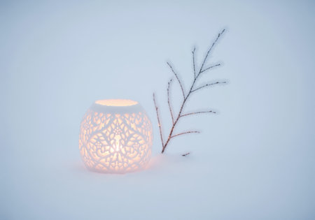 The intricate lace pattern of a glowing white ceramic candle holder casts a warm light on the surrounding fresh snow, symbolizing cozy winter holidays.の素材