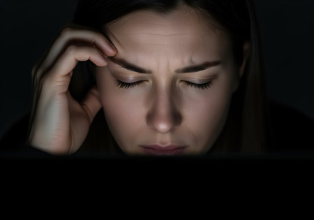 A dramatic close up shot of a young woman experiencing intense stress and headache, illuminated only by the glow of a computer screen while working late.の素材