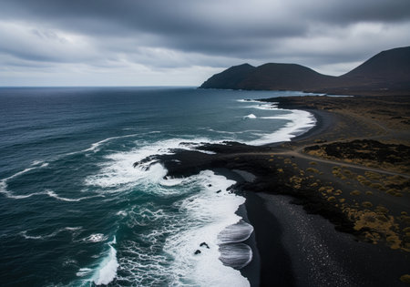 High angle view captures the dramatic contrast between the dark turquoise ocean and the black volcanic sand beach. waves crash powerfully onto the rugged shore under a moody, overcast sky.の素材