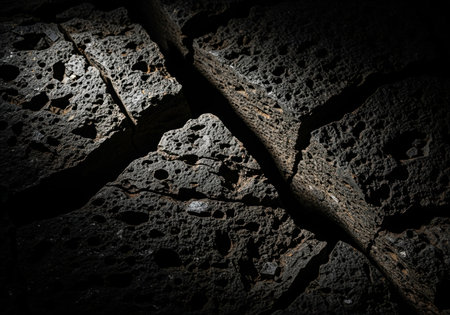 A dramatic, low key macro shot highlighting the rough, porous texture of dark volcanic basalt rock, featuring deep cracks and fissures.の素材