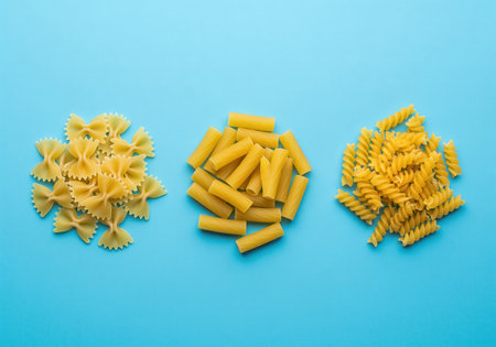 Three small piles of dry, uncooked italian pasta shapes farfalle, rigatoni, and fusilli arranged horizontally on a vibrant blue surface, top view.の素材