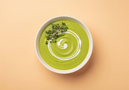 Vibrant green broccoli cream soup served in a white bowl, garnished with a decorative cream swirl and fresh microgreens, presented on a light peach background. healthy, vegan, and gourmet appetizer.の素材