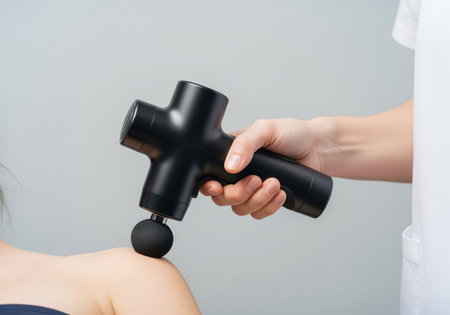 Percussion massage gun, held by a therapist hand, applying deep tissue massage to a patient shoulder for pain relief and muscle recovery in a clinical setting.の素材